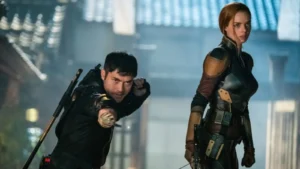 Two characters in tactical combat gear from the film Snake Eyes stand ready for action, one aiming forward in a fighting stance while the other looks alert beside him in a dimly lit urban setting