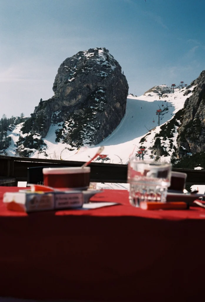 Table with espresso cups, cigarette pack, and glass in the foreground overlooking a snowy ski slope and dramatic Dolomite rock formation in Cortina d’Ampezzo