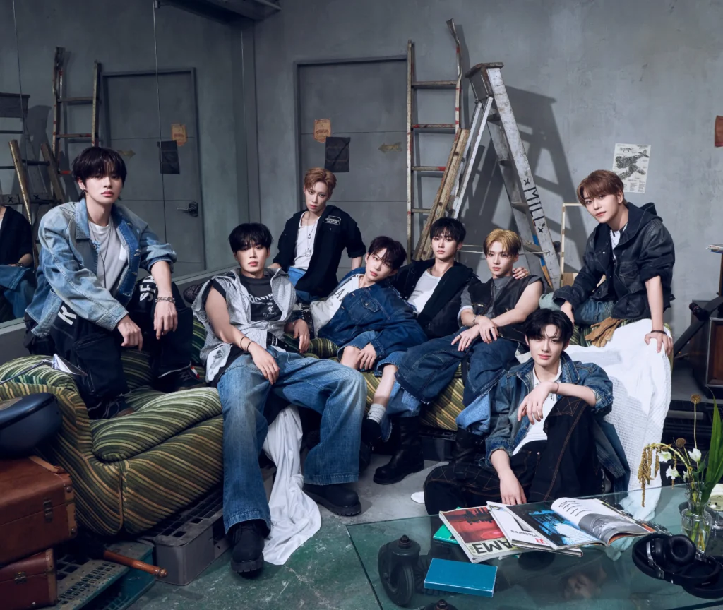 Alpha Drive One (ALD1) members posed together in an industrial studio setting for their Euphoria debut era photoshoot, wearing layered denim and contemporary streetwear while seated across a vintage sofa surrounded by ladders, magazines, and studio props