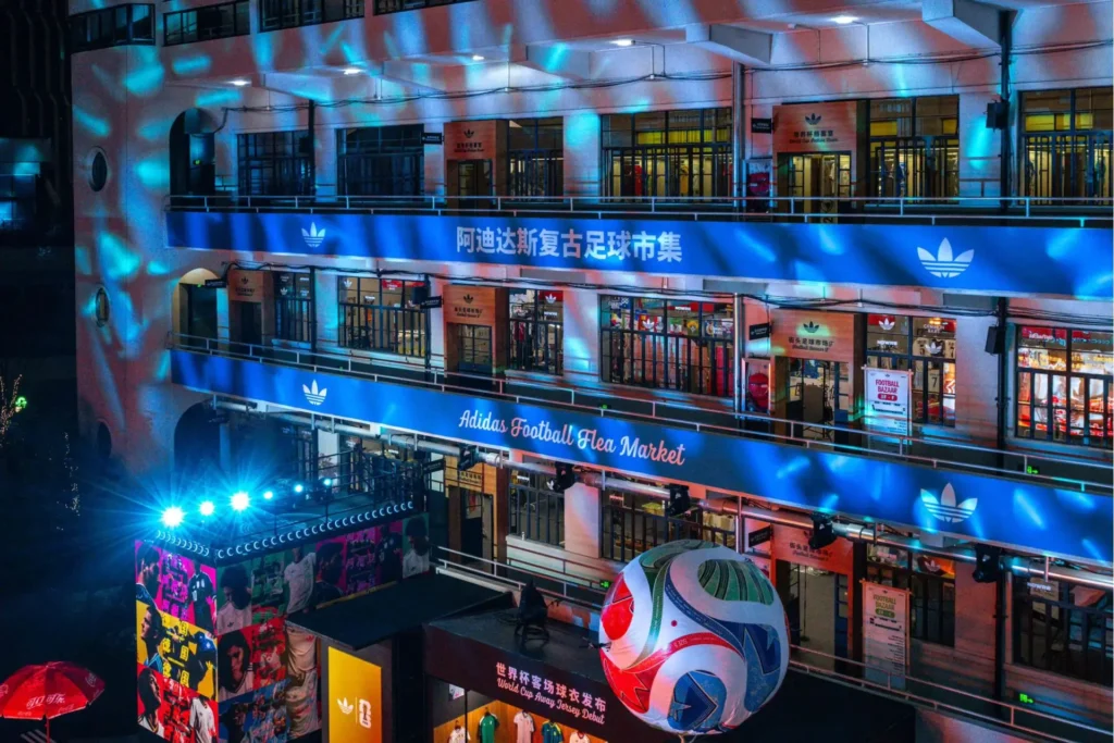 Exterior night view of adidas’ Shanghai football flea market activation, featuring a multi-level building illuminated in blue light with large “Adidas Football Flea Market” signage and immersive event installations below