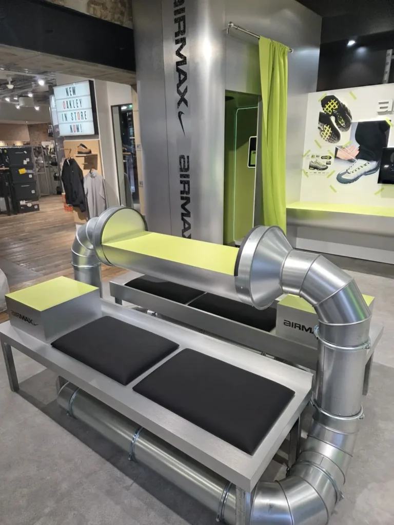 Industrial-inspired in-store installation featuring a brushed metal bench constructed from exposed ventilation ducting, accented with neon lime surfaces and black seat pads, positioned within a retail environment branded with Nike Air Max signage and product displays, evoking a fusion of technical infrastructure and sneaker innovation
