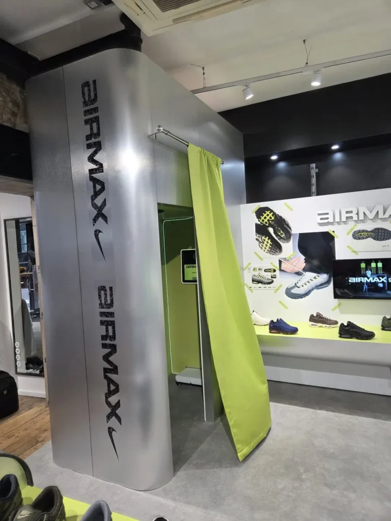 Futuristic in-store installation featuring a metallic changing-room structure branded with Nike Air Max typography, accented by a lime green curtain and industrial finishes, set within a sneaker retail space displaying Air Max footwear and campaign visuals, merging utilitarian design with performance-driven aesthetics