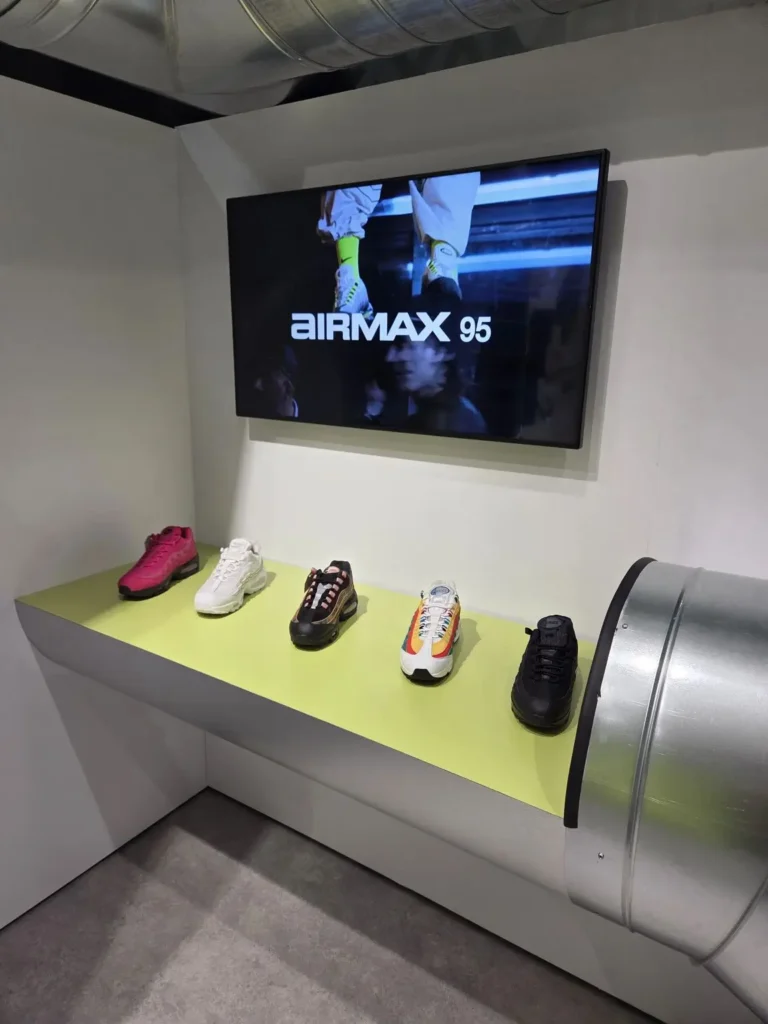 Curated retail display featuring multiple Nike Air Max 95 colorways arranged on a lime green platform beneath a wall-mounted screen playing Air Max campaign footage, framed by exposed metallic ductwork and clean architectural lines, presenting the silhouette within a refined, industrial-inspired store environment