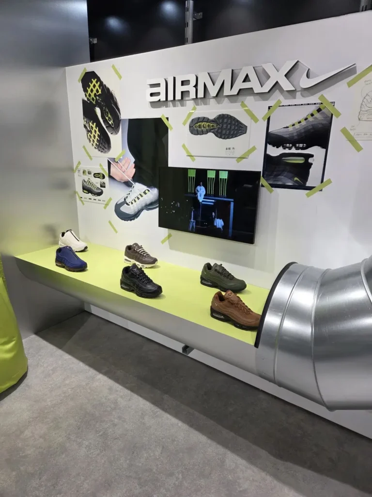 Wall-mounted Nike Air Max installation featuring campaign imagery, technical sole diagrams, and a digital display, paired with a lineup of Air Max sneakers arranged on a lime green platform, all set within an industrial-inspired retail environment accented by exposed metallic ductwork and clean architectural surfaces