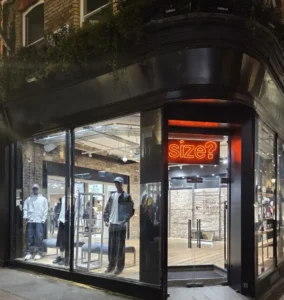 Exterior view of a size? retail store on a city corner, featuring a glowing orange neon logo above the entrance, large glass windows displaying mannequins in streetwear, and an industrial-style interior with exposed brick and lighting, presenting a refined, urban sneaker and apparel retail environment at night