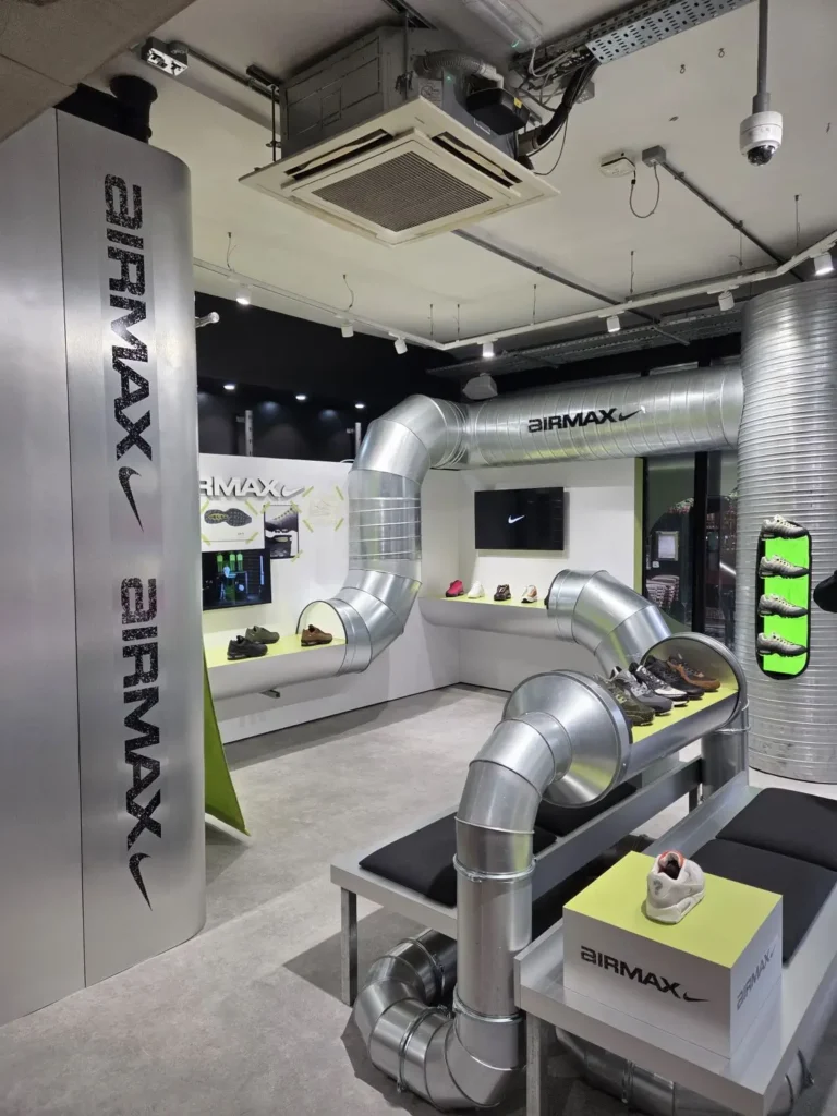 Wide interior view of a Nike Air Max retail installation featuring exposed metallic ductwork integrated into seating and display structures, lime green platforms showcasing Air Max sneakers, and clean architectural surfaces, creating an industrial, performance-driven environment that visually echoes the concept of airflow and cushioning technology