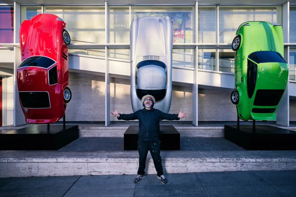 Mr Brainwash posing with arms open in front of three vertically mounted cars—red, silver, and green—outside the Mr Brainwash Art Museum in Beverly Hills
