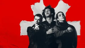 Early lineup of the Red Hot Chili Peppers featuring Anthony Kiedis, Flea, and Hillel Slovak posed against a bold red background with the band’s iconic asterisk symbol, evoking the group’s raw beginnings and enduring brotherhood
