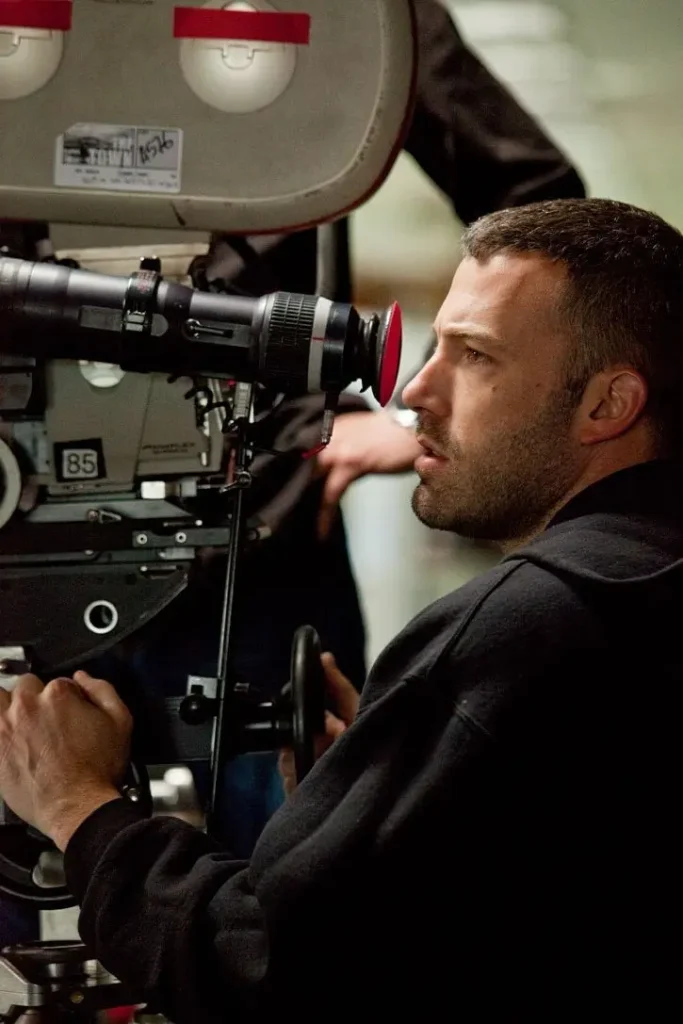Ben Affleck operating a professional motion picture camera on a film set, focusing through the viewfinder while directing a scene during production