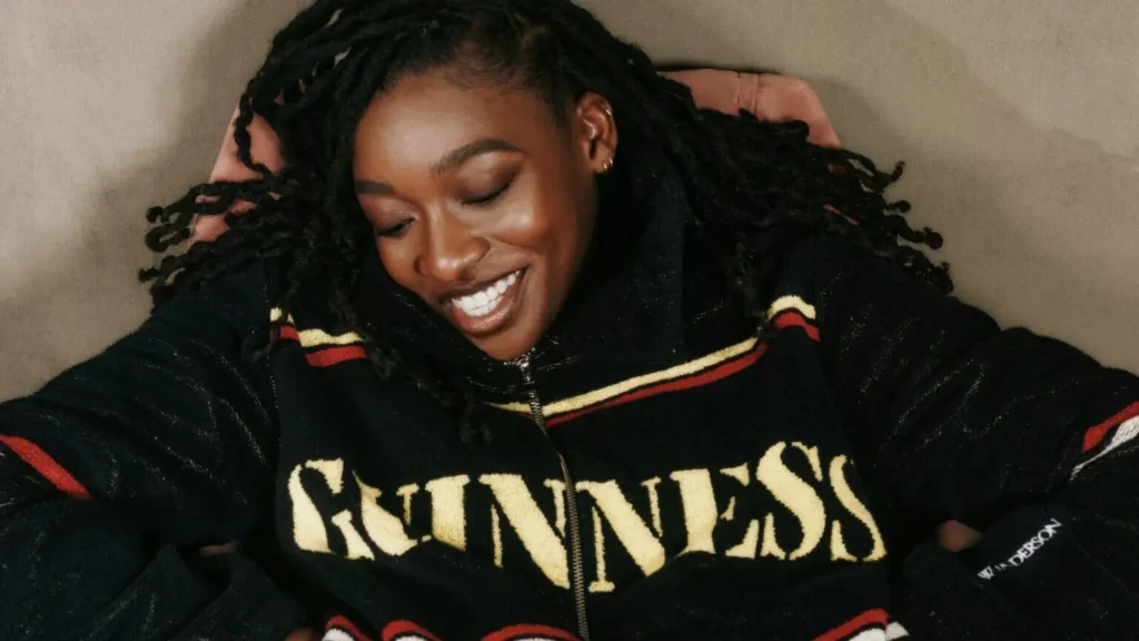 Little Simz smiling in JW Anderson x Guinness black zip jacket with bold Guinness logo