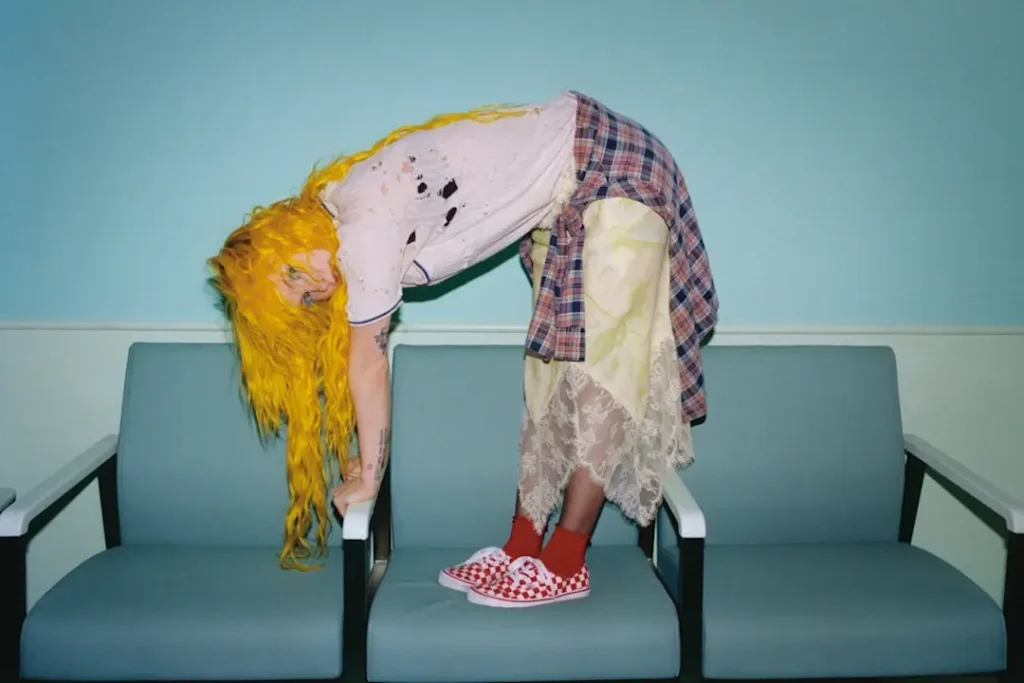 Hayley Williams bends across a row of waiting-room chairs with bright yellow hair cascading downward, wearing red checkerboard Vans Authentic sneakers as part of the Vans “Off The Wall” 60th anniversary campaign