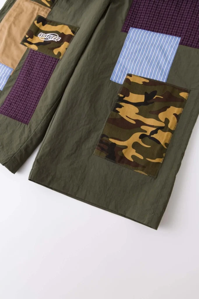 Lower section detail of SSSTUFF patchwork shorts showing large camouflage panel layered with blue striped fabric and adjacent burgundy grid textile on olive base