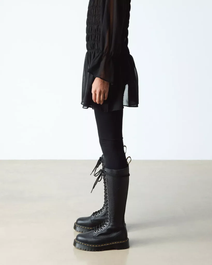 Model wearing Dr. Martens 1B60 Bex knee-high black leather boots styled with a black dress and tights, showcasing full-length silhouette