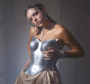 Portrait of the artist googly eyes wearing a sculptural silver metallic corset and layered chain jewelry with a jeweled headpiece, posing in soft studio lighting