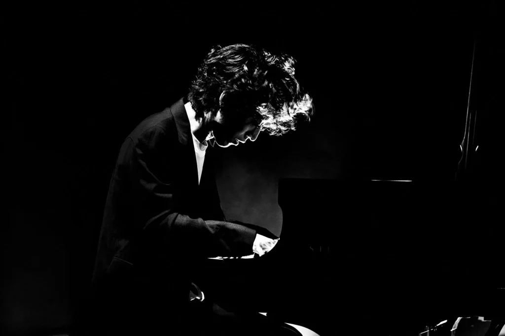 Black-and-white portrait of Hayato Sumino seated at a grand piano, captured in a moment of intense focus, with dramatic lighting emphasizing his silhouette and expressive posture during performance