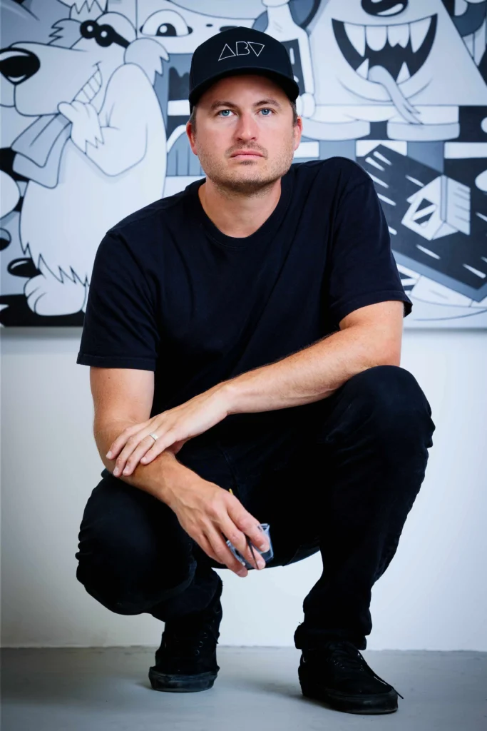 Greg Mike crouching in his studio wearing a black cap and T-shirt, posed in front of a black-and-white mural featuring his signature cartoon-style characters