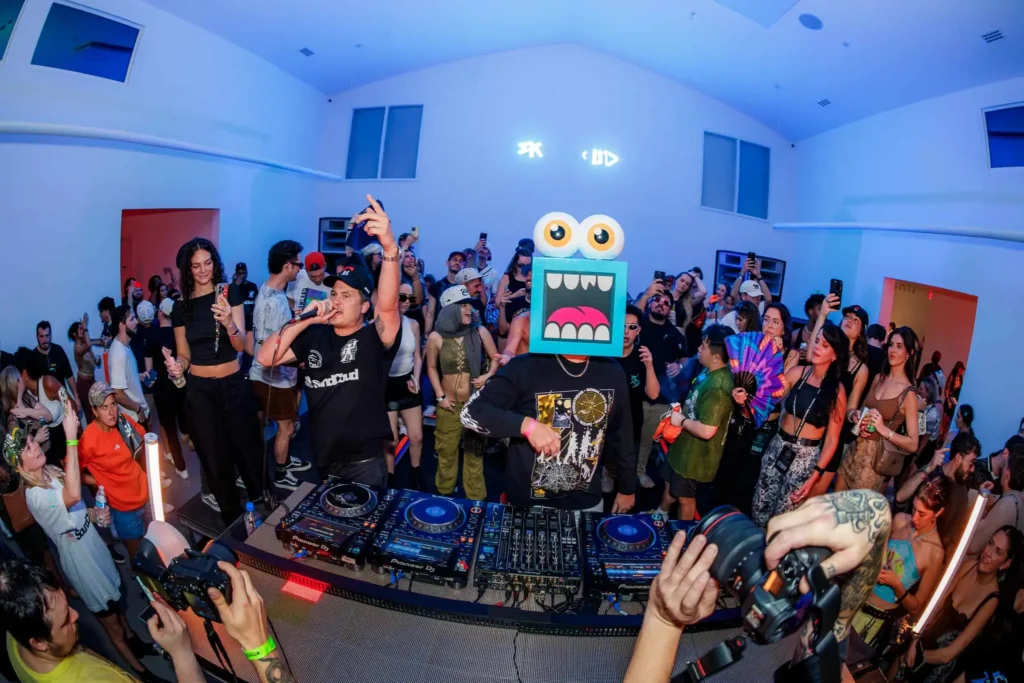 Crowded DJ set during a Greg Mike event with a performer wearing a Loudmouf character head while music plays to a packed audience inside a gallery space