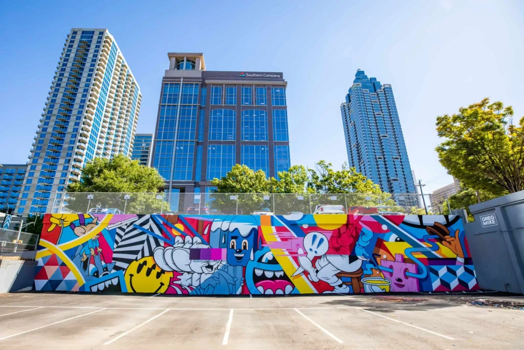 Large-scale Greg Mike mural featuring colorful Loudmouf characters and geometric patterns painted along a wall beneath Atlanta skyscrapers