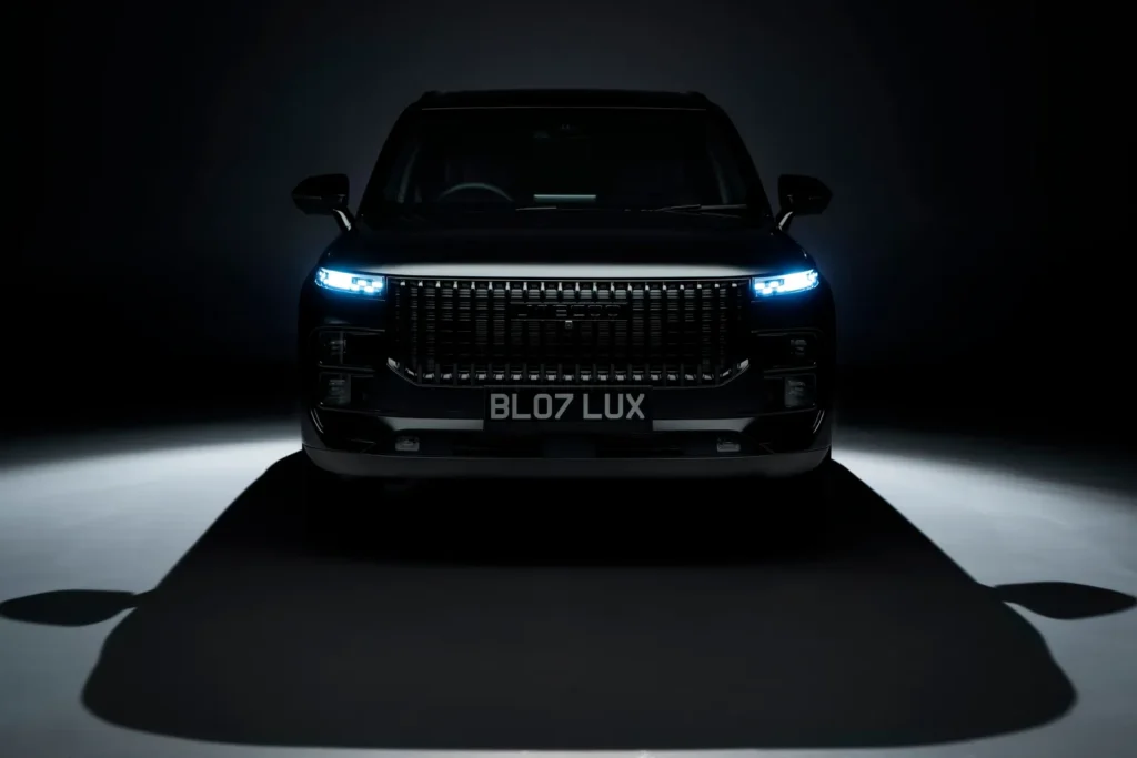 Front-facing studio shot of the Jaecoo 7 SUV in a dark setting, with illuminated blue LED headlights, a wide vertical-slat grille, and a centered “BL07 LUX” plate casting a sharp shadow on the floor