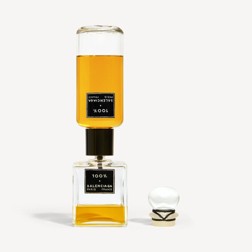 Balenciaga 100% perfume bottle with amber liquid stacked beneath a second bottle, with the glass stopper placed beside the base on a neutral background