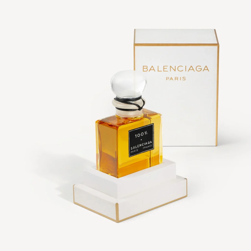 Balenciaga 100% perfume bottle with amber fragrance displayed on a white pedestal beside its white and gold Balenciaga Paris box packaging
