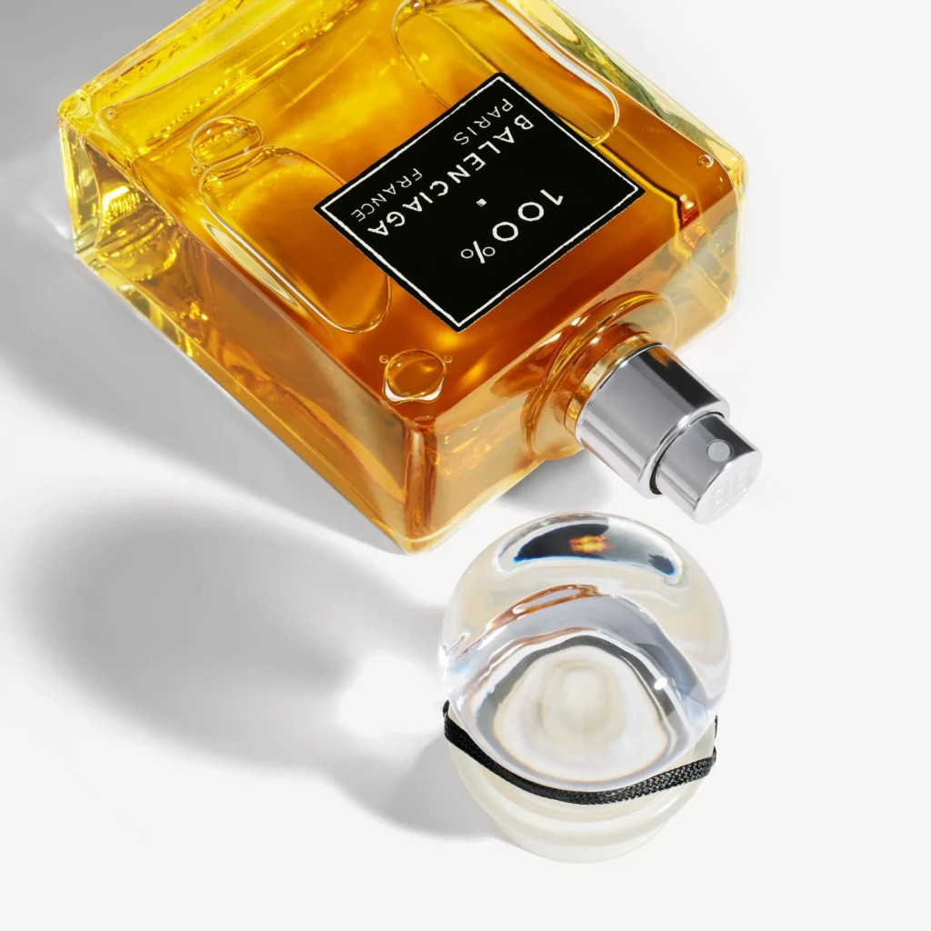 Balenciaga 100% perfume bottle lying on its side showing the spray atomizer, with amber fragrance inside a square glass bottle and the round glass stopper placed beside it