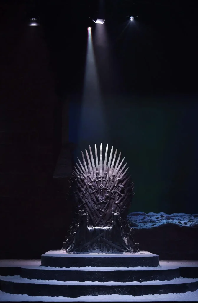 Iron Throne illuminated by a single spotlight in a dark hall, the iconic seat of power from Game of Thrones standing atop snow-covered steps surrounded by silhouette