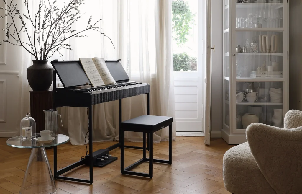 Donner OURA Digital Piano with matching bench placed in a bright, minimalist living room beside tall windows and sheer curtains, featuring an open sheet music book on the stand and modern home décor