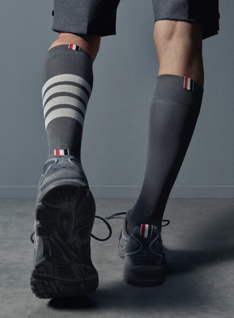 Rear lifestyle view of Thom Browne x ASICS GEL-Kayano 14 sneakers worn with tailored shorts and grey knee-high socks featuring signature four-bar stripes and tricolor tabs, highlighting outsole traction and heel branding in a minimal studio setting