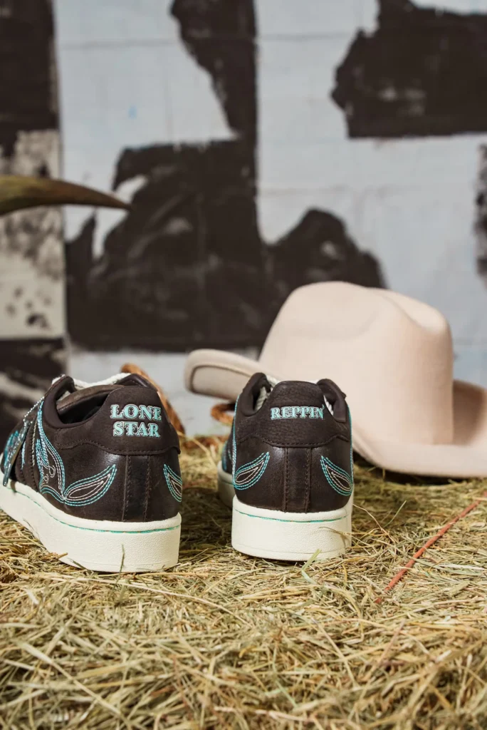 Rear view of adidas Superstar “Lone Star Reppin’” sneakers with turquoise embroidery and “Lone Star” and “Reppin’” heel text, placed on hay beside a cowboy hat