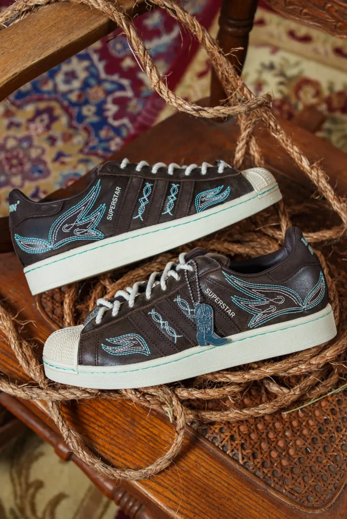 Pair of adidas Superstar “Lone Star Reppin’” brown leather sneakers with turquoise Western embroidery and cowboy boot lace charm, styled with rope on a wooden chair