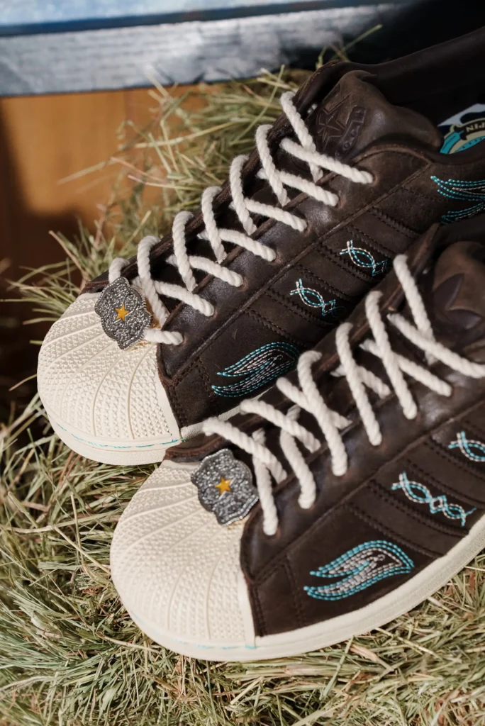 Top view of adidas Superstar “Lone Star Reppin’” brown leather sneakers with turquoise Western embroidery, shell toe, and star badge lace charm resting on hay