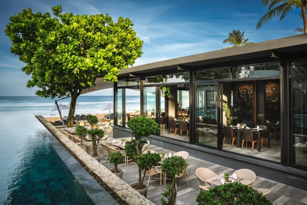Exterior view of The SEASONS at Aleenta Phuket-Phang Nga, with a glass-walled dining space beside an infinity pool overlooking the Andaman Sea, framed by a large tropical tree and beachfront terrace seating under a clear blue sky