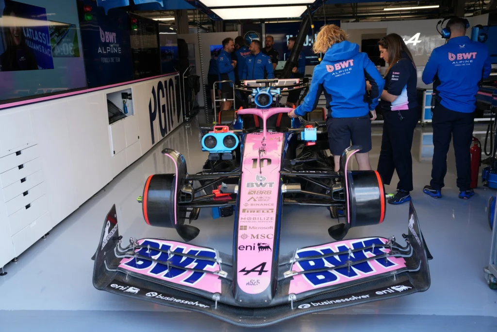 Front view of a BWT Alpine Formula 1 car inside the team garage, surrounded by crew members in blue uniforms preparing and inspecting the vehicle