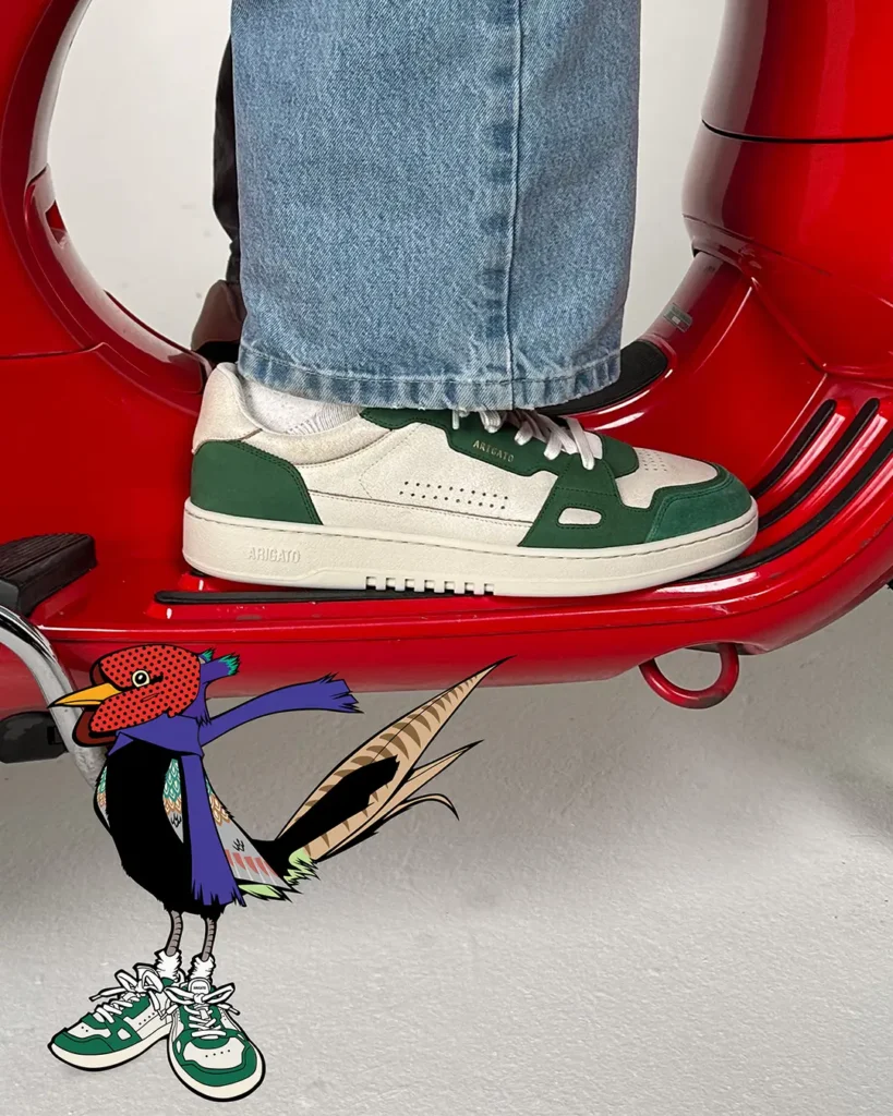 Axel Arigato green-and-cream shoe worn on a red scooter step with illustrated bird mascot from the Akaia collab with Reggieknow