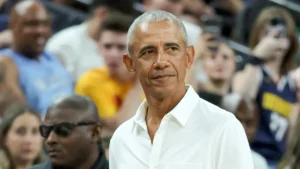 Barack Obama courtside at an NBA game, wearing a white shirt and observing the action with a composed expression