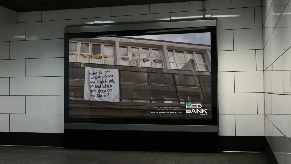 Public billboard display showing a Bed Bank campaign message on a building façade reading “How do you wake up on the right side of bed when you sleep on the floor?”, raising awareness of bed poverty in an urban setting