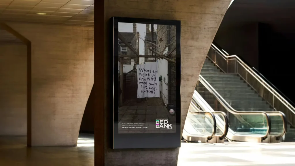 Digital advertising display in a public transit space showing a Bed Bank campaign poster with the message “What’s the point in dreaming when you’re not even sleeping?”, highlighting the reality of bed poverty