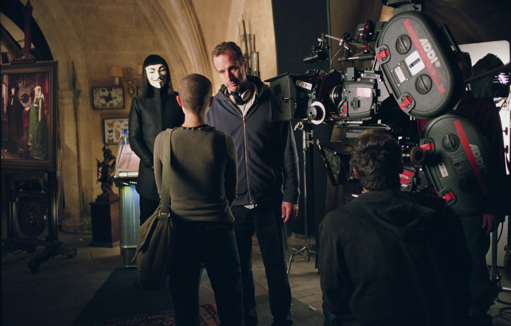 Behind-the-scenes on the set of V for Vendetta, with James McTeigue directing Natalie Portman during a pivotal scene, while Hugo Weaving stands in costume as V in the background beside the production camera setup
