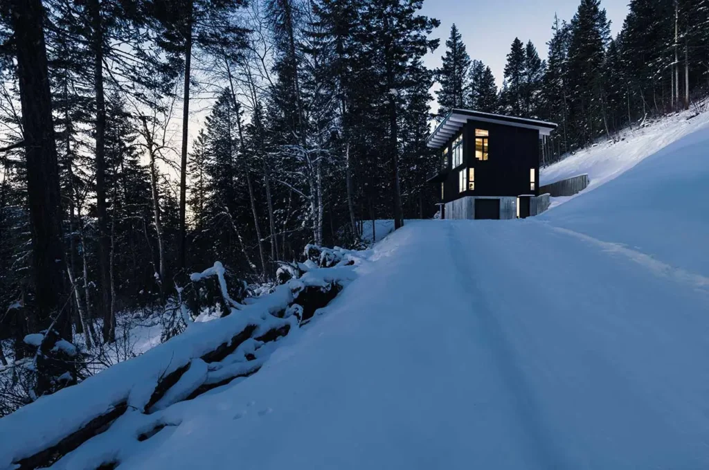 Columbia River Valley Lookout cabin by Twobytwo Architecture glowing at dusk on a snowy hillside surrounded by dense pine forest near Golden, British Columbia