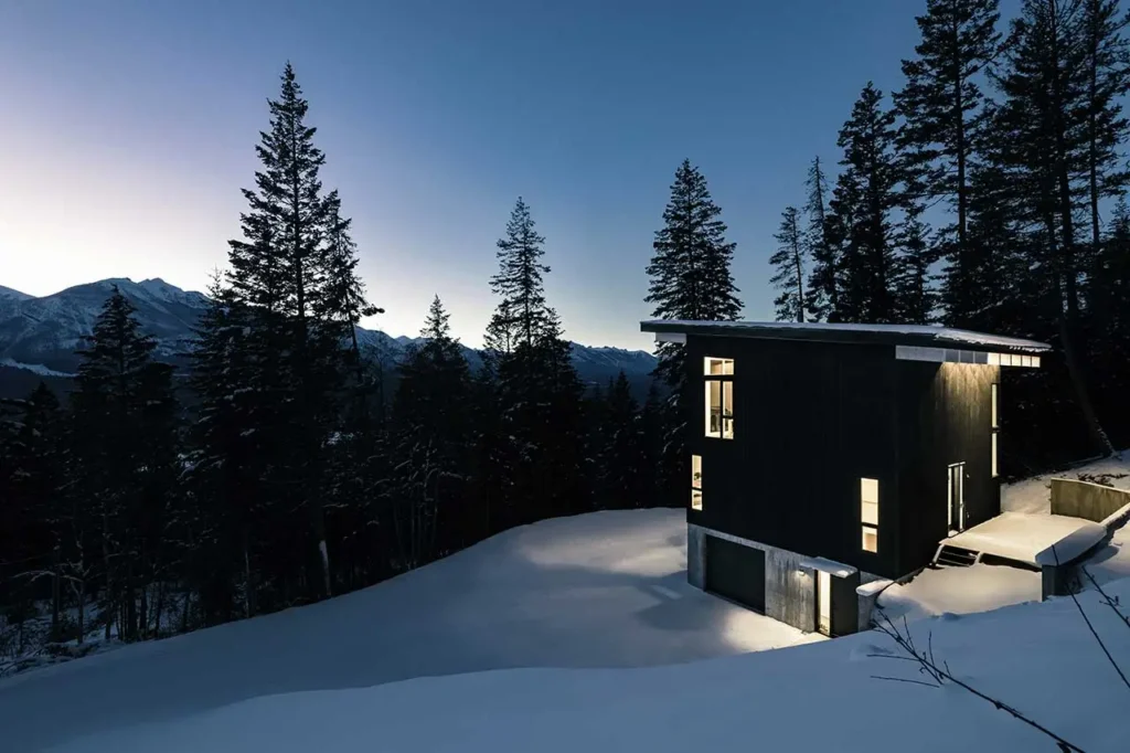 Columbia River Valley Lookout cabin by Twobytwo Architecture illuminated at twilight on a snowy hillside overlooking the forested Columbia River Valley near Golden, British Columbia