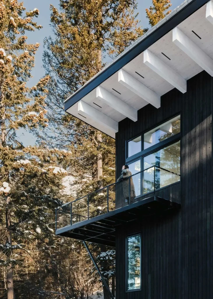 Cantilevered balcony of the Columbia River Valley Lookout cabin by Twobytwo Architecture overlooking a snowy pine forest in Golden, British Columbia, with black timber siding and a white overhanging roof