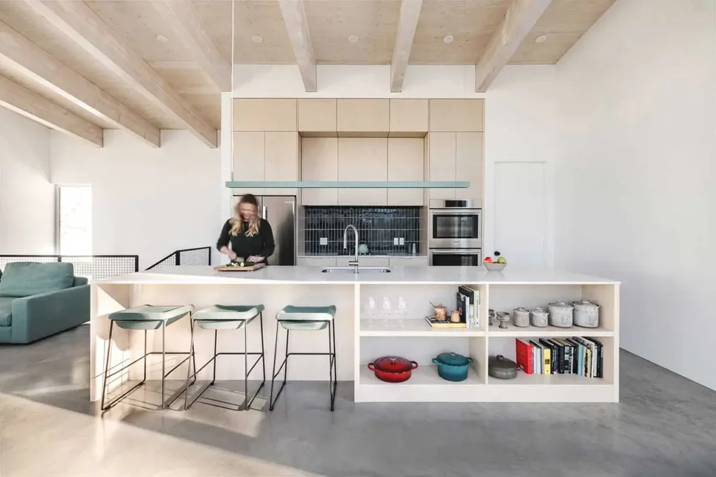 Minimalist kitchen interior inside the Columbia River Valley Lookout cabin by Twobytwo Architecture featuring a large white island, light wood ceiling beams, built-in shelving, and modern appliances