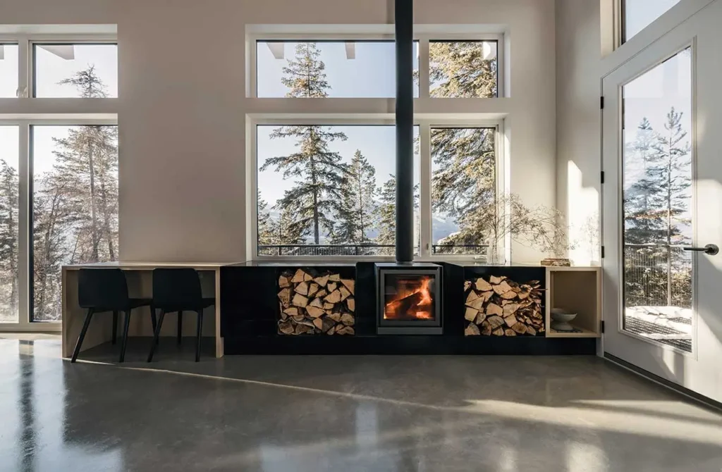 Living room interior of the Columbia River Valley Lookout cabin by Twobytwo Architecture featuring a wood-burning stove, stacked firewood storage, and large windows overlooking snowy pine forest in Golden, British Columbia