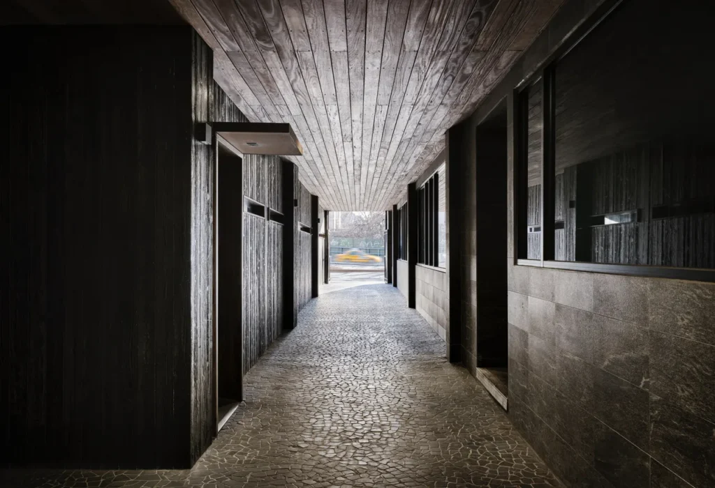 A narrow, atmospheric corridor with a textured stone floor, dark wood-paneled walls, and a slatted wood ceiling, leading toward daylight and a blurred yellow taxi passing outside