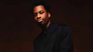 Portrait of rapper Denzel Curry wearing a dark textured shirt against a warm brown studio background, looking directly at the camera