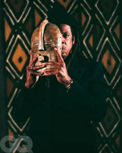 A sharply lit portrait of a man holding a carved wooden African mask over half of his face, revealing one eye and part of his expression, while wearing a dark coat and knit cap against a patterned geometric backdrop, emphasizing themes of duality, identity, and concealment