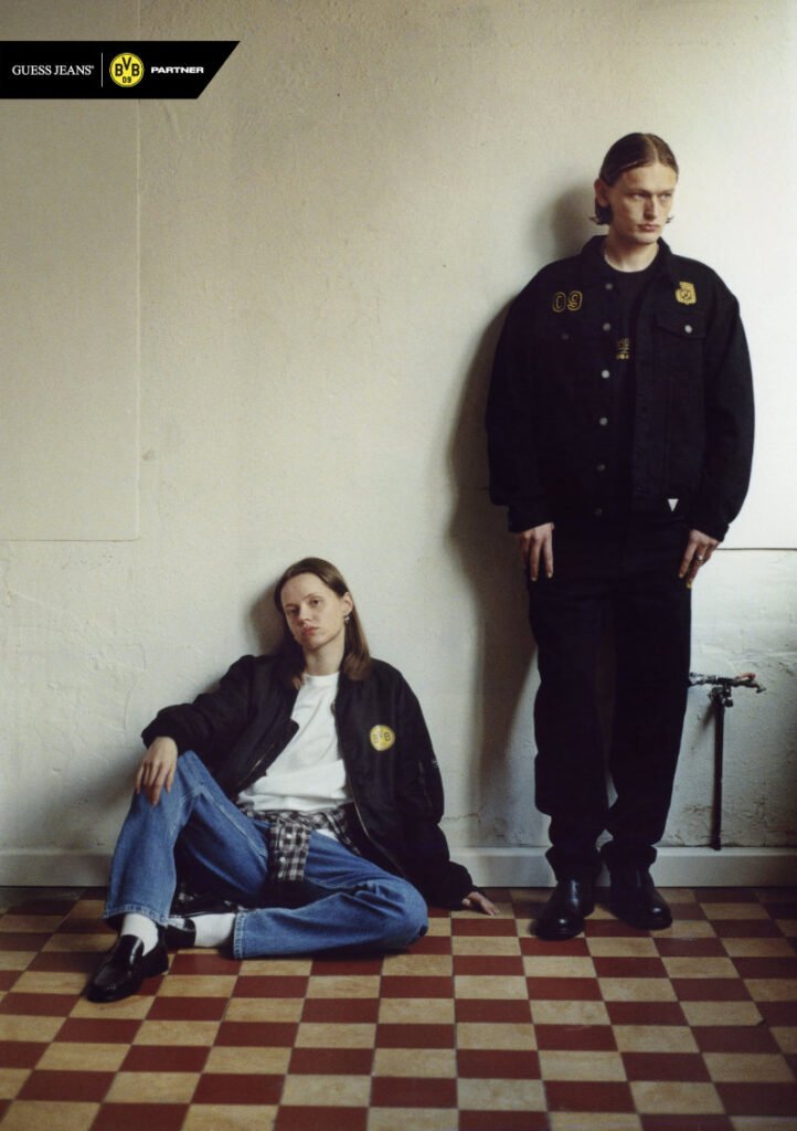 Two supporters wearing black GUESS JEANS x Borussia Dortmund jackets with BVB 09 patches pose indoors on a checkered floor, highlighting the capsule’s football-inspired streetwear aesthetic