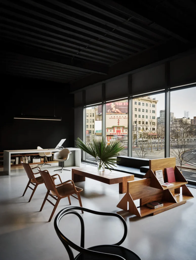 A refined studio lounge with mid-century wooden chairs, a low table with a plant, and a sculptural wood display shelf, set beside floor-to-ceiling windows overlooking a city street, blending workspace and living-room comfort