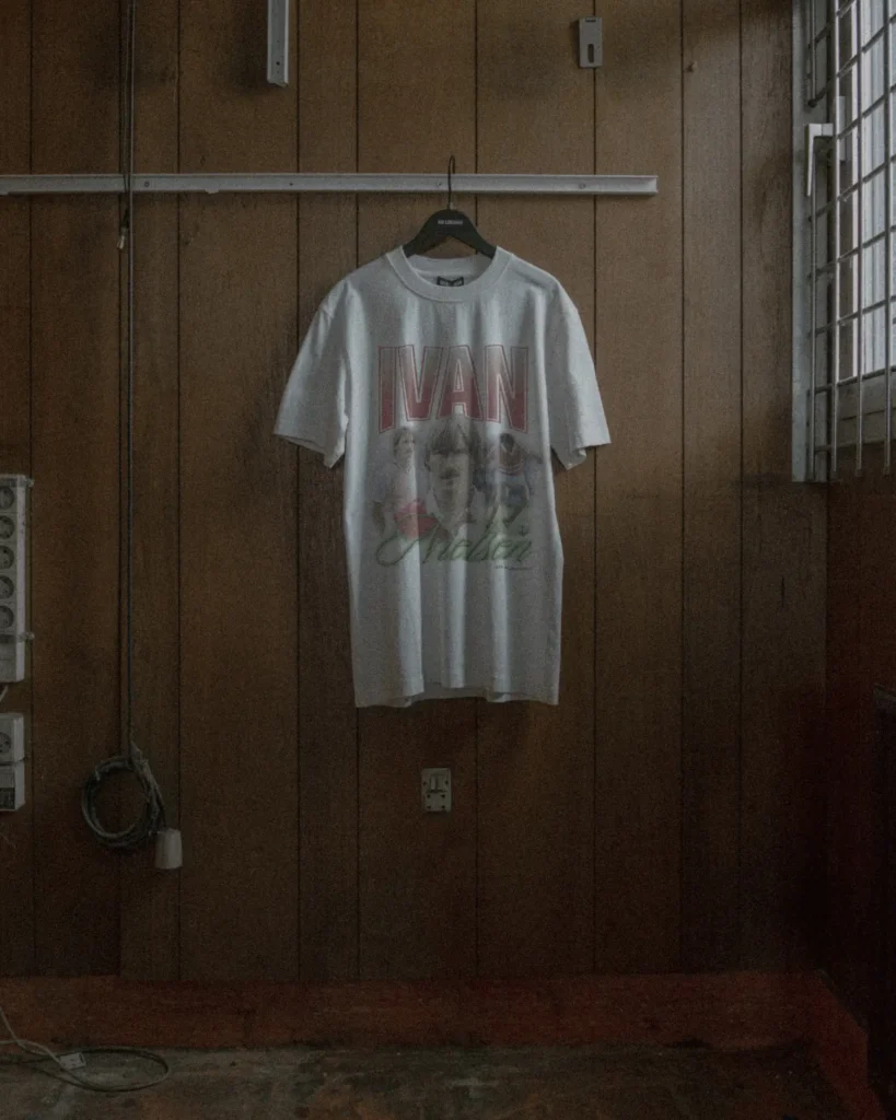 A graphic T-shirt hangs on a black hanger against a wood-paneled wall, featuring a faded portrait print and bold “IVAN” lettering, referencing football iconography within Han Kjøbenhavn’s Pre-AW26 collection